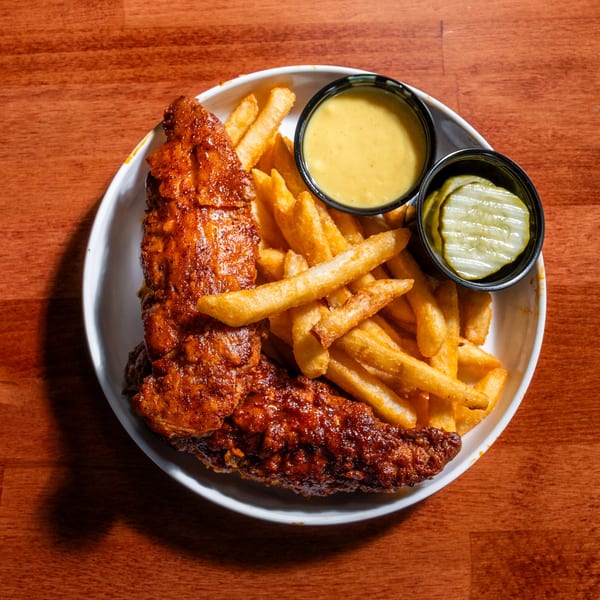 Spicy chicken tenders with french fries, a side of sauce, and pickles on a white plate.