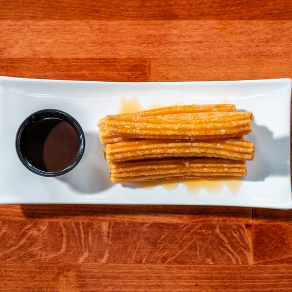 Churros drizzled with syrup and a side of chocolate dipping sauce on a white rectangular plate.