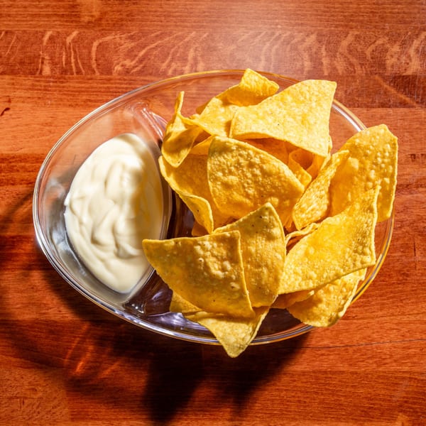 A glass bowl of crispy tortilla chips and creamy white dip on a wooden table.