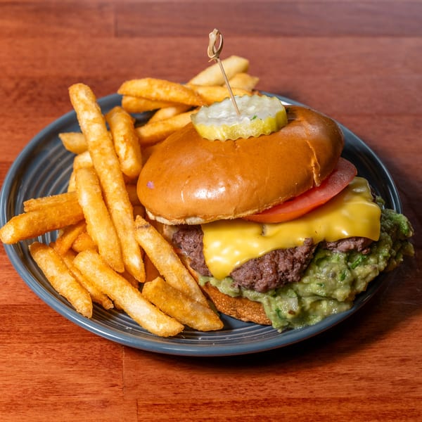 A cheeseburger with guacamole, tomato, and a pickle spear, served with a side of french fries.