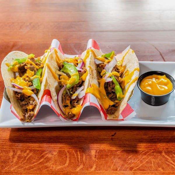 Three Al Tacos Locos tacos with meat, cheese sauce, onions, and green peppers on a white plate.