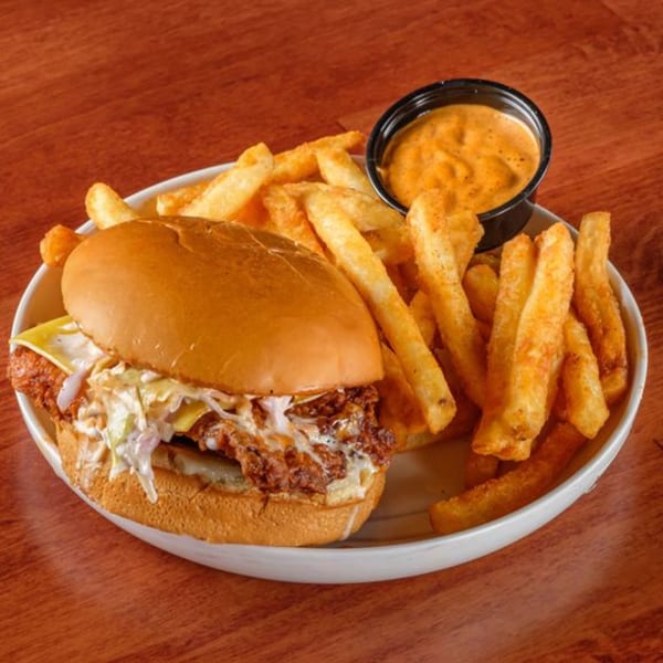 A crispy chicken sandwich with cheese and coleslaw, served with a side of fries and dipping sauce.